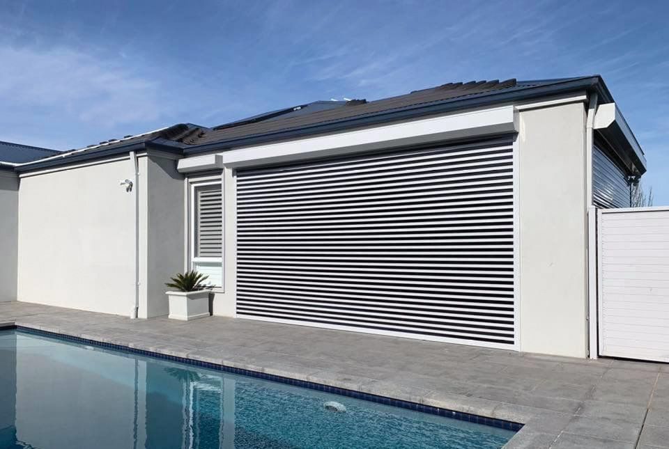 A modern home with a pool, featuring a closed, striped security shutter on the garage and bright blue sky.