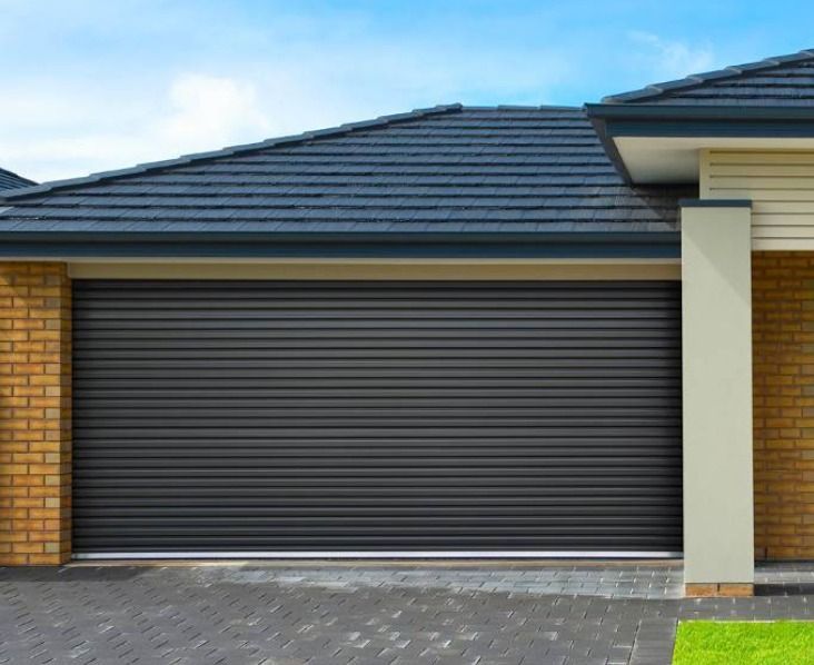 Dark gray roll-up garage door with brick and beige exterior against a blue sky.