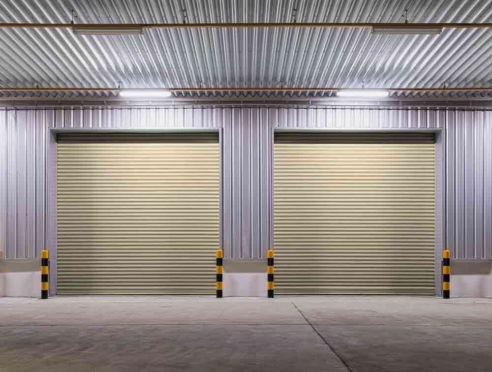 Two beige roller doors in a metal warehouse, with black and yellow bollards.
