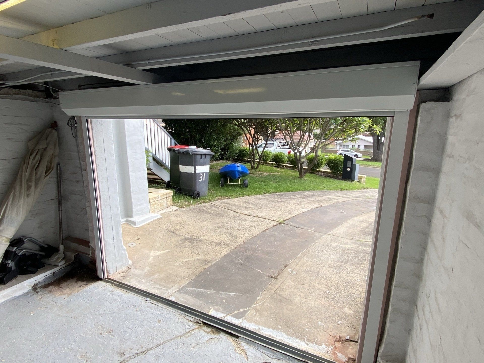 Garage door open, revealing a driveway and yard. Gray door, concrete and greenery visible.