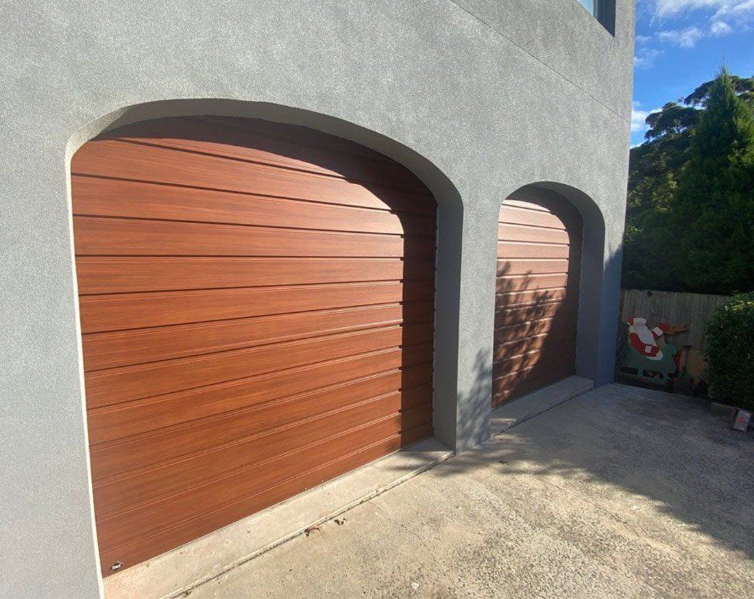 Two brown arched garage doors set into a gray stucco wall, in a driveway.