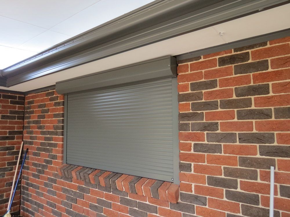 Gray roll-down security shutter on a brick wall beneath a gray eave.