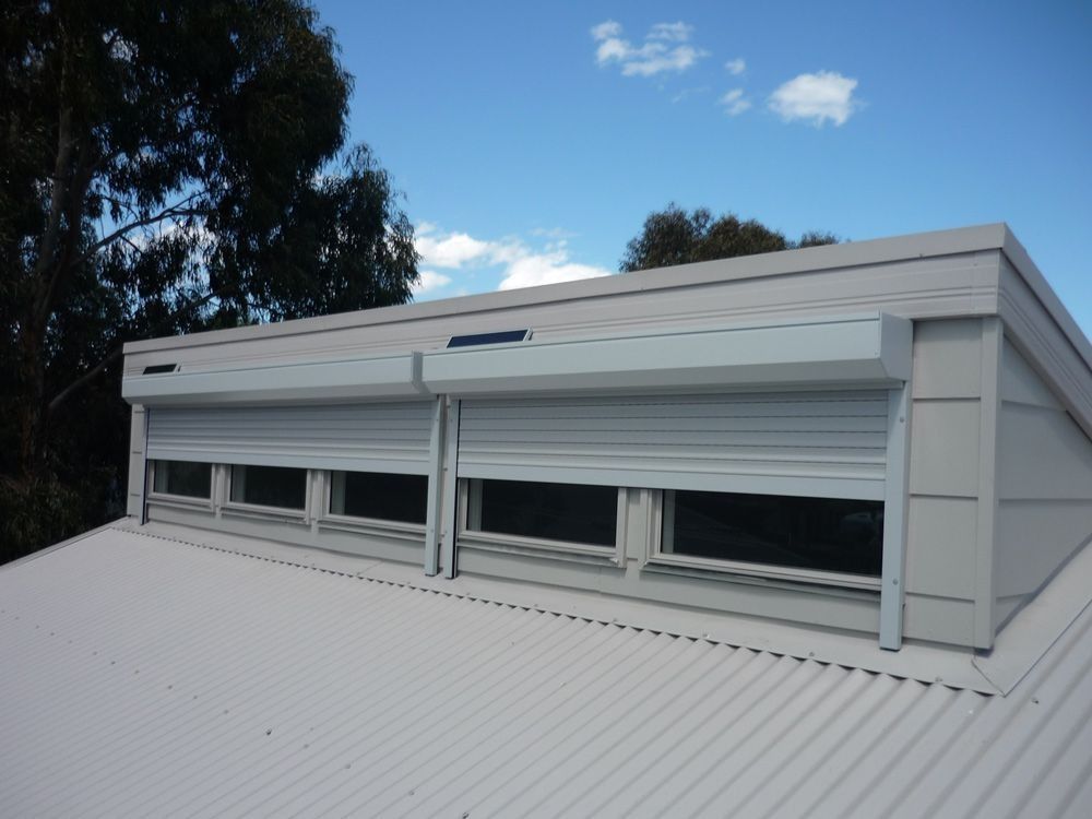 White roller shutters covering windows on a white roof, under a blue sky.