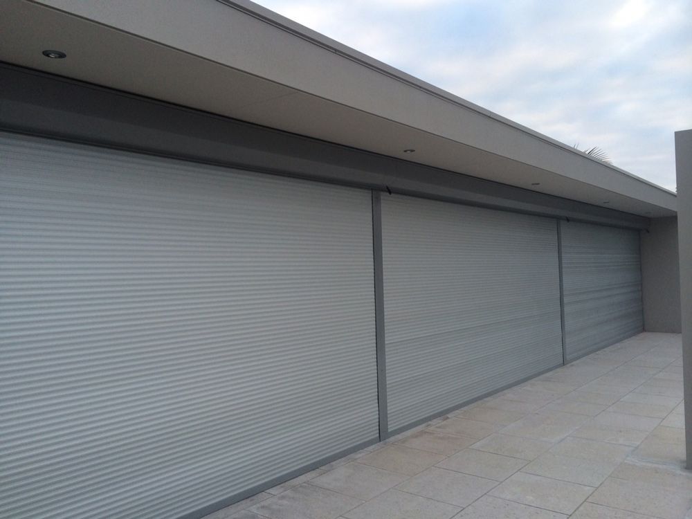 Exterior view of building with three closed, gray, roller shutters and a stone floor.