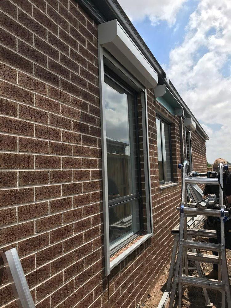 Brick building exterior with a window and installed white roller shutter. A ladder and equipment are on the right.