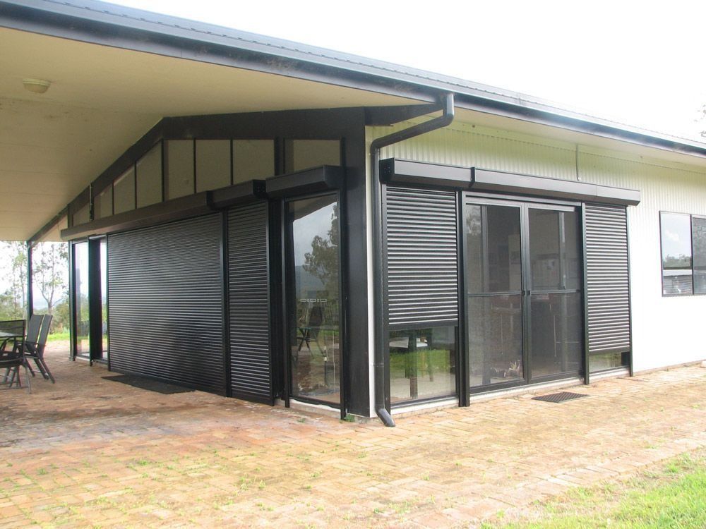 Black roller shutters on an outdoor patio of a white house. A dining set is in the shade.