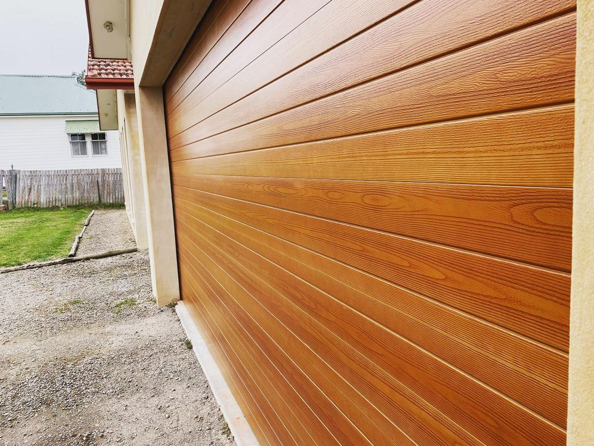 Wood-grained garage door, tan walls, gravel driveway, and a glimpse of a house with a green lawn.