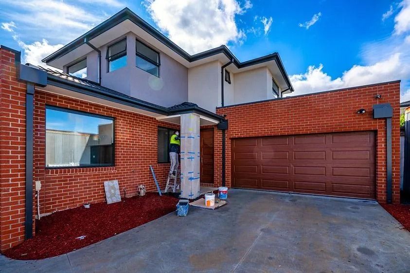 Two-story brick house with brown garage door, blue sky, and red landscaping. Construction work in progress.