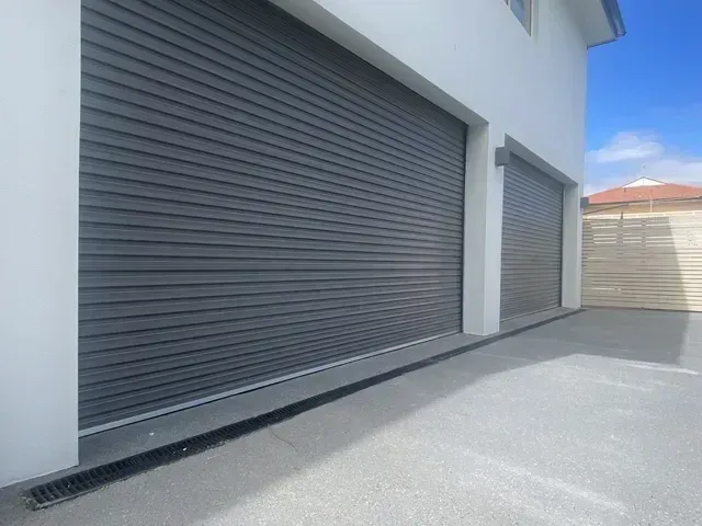 Gray rolling garage doors on a white building, gray concrete driveway, sunny day.