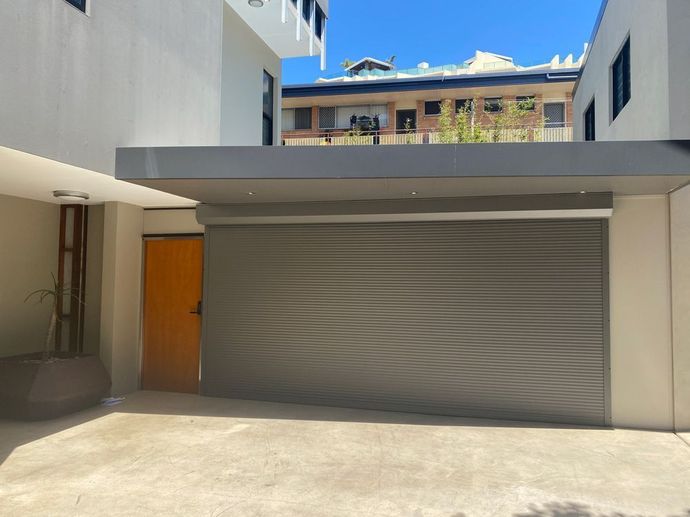 Gray roller shutter covering an opening, next to a brown door in a light-colored building courtyard.
