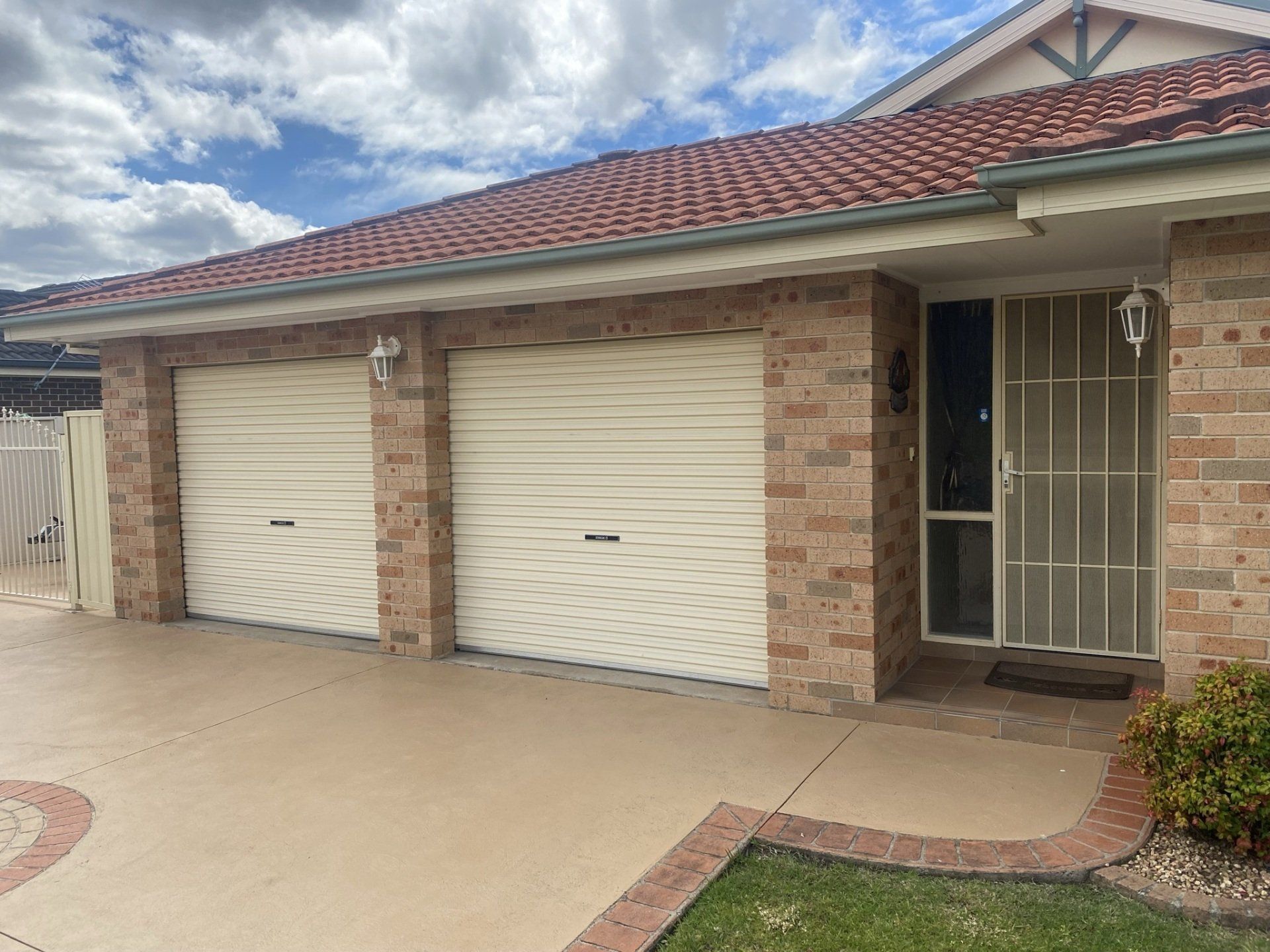 Two beige garage doors and a front door with security screen on a brick house, under a tiled roof.