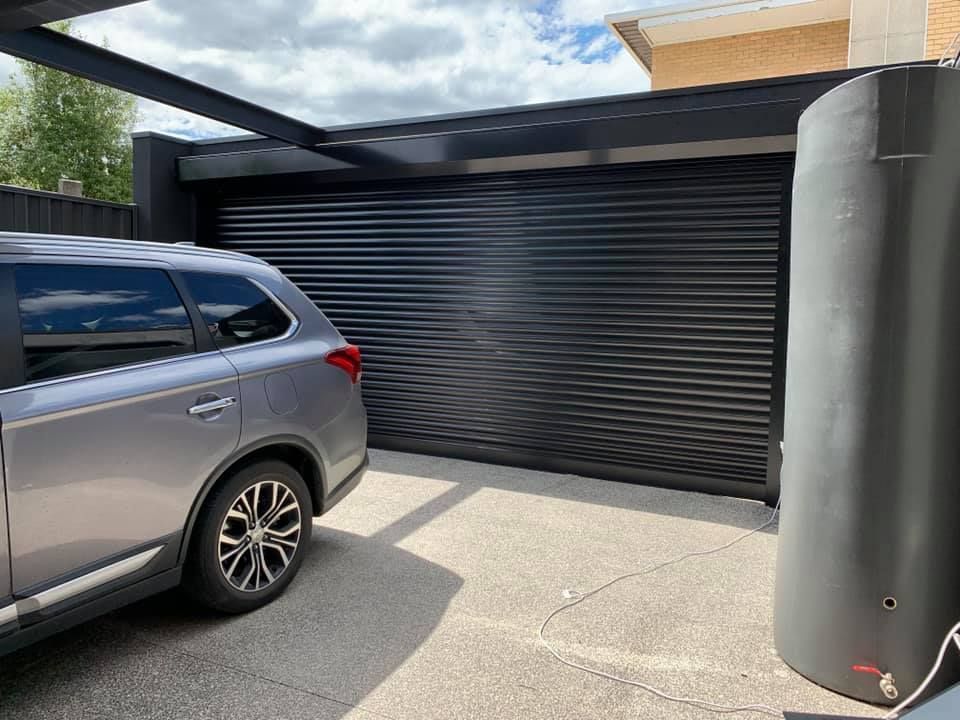 Silver SUV parked under a black carport next to a black corrugated garage door and a water tank.