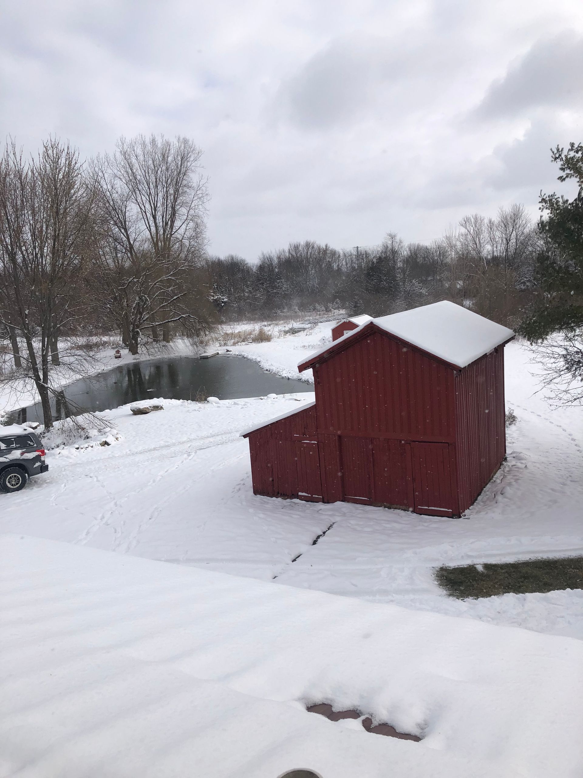 A red barn is covered in snow next to a pond.