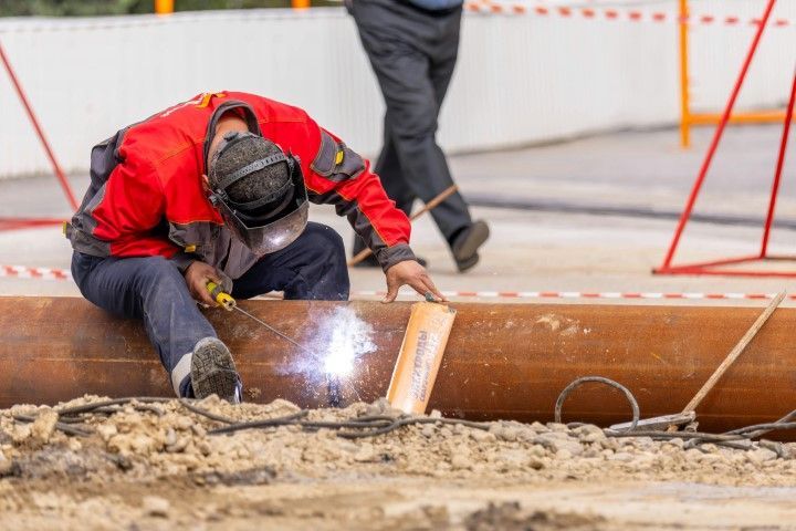A welder in a red jacket and protective gear welds a large metal pipe on a Newton construction site.