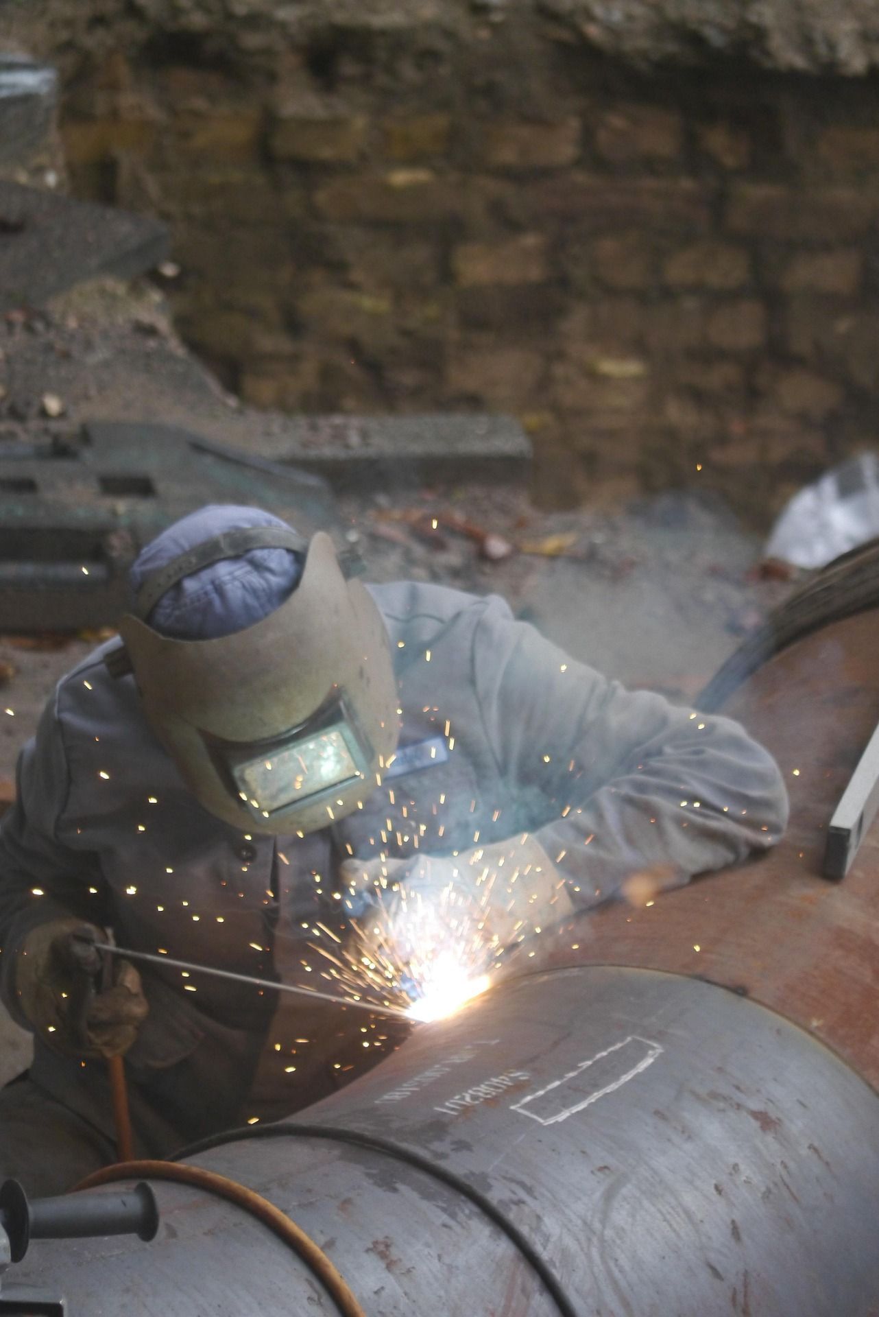 A welder in protective gear and a helmet uses a welding torch on a large metal pipe, creating bright sparks.