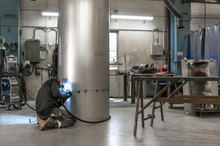 A person kneeling in a workshop, welding a seam on a large, vertical stainless steel cylindrical tank.