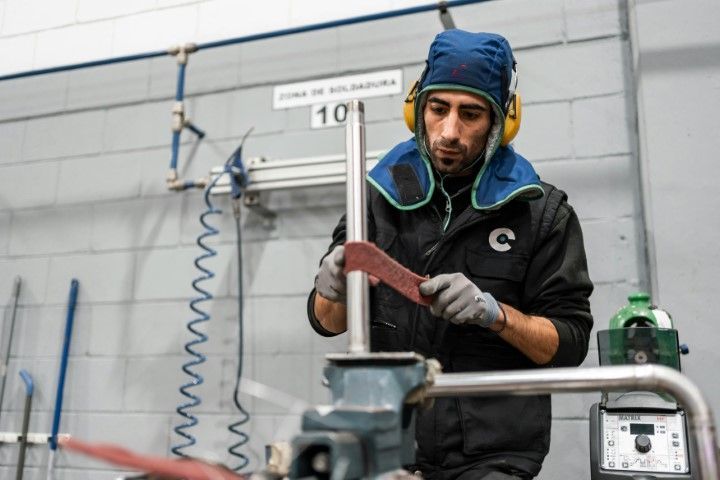 A person in a welding shop wearing protective gear, sanding a metal rod secured in a vice.