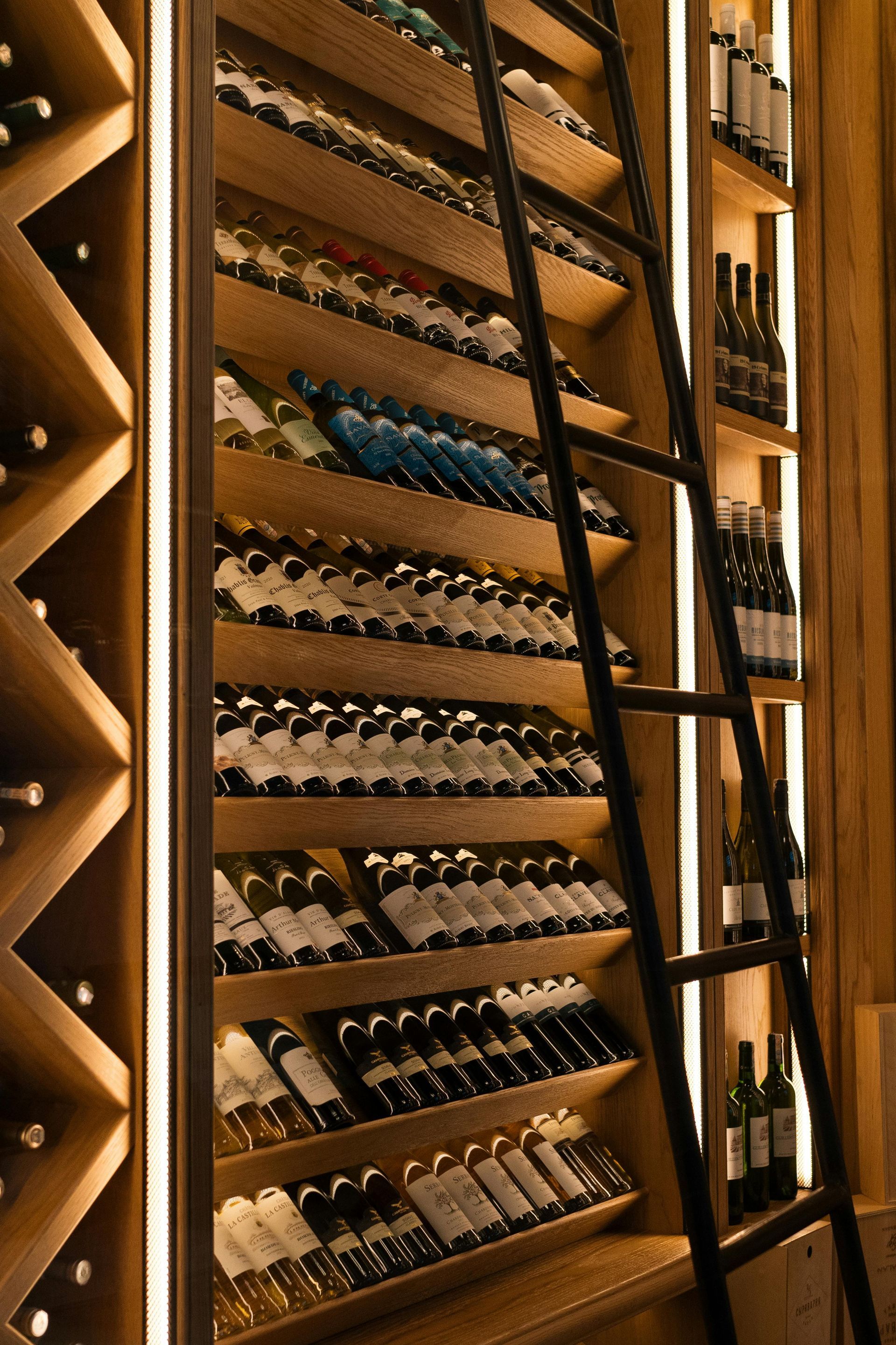Wooden wine rack filled with bottles, lit with vertical lights, a black ladder leans against it.