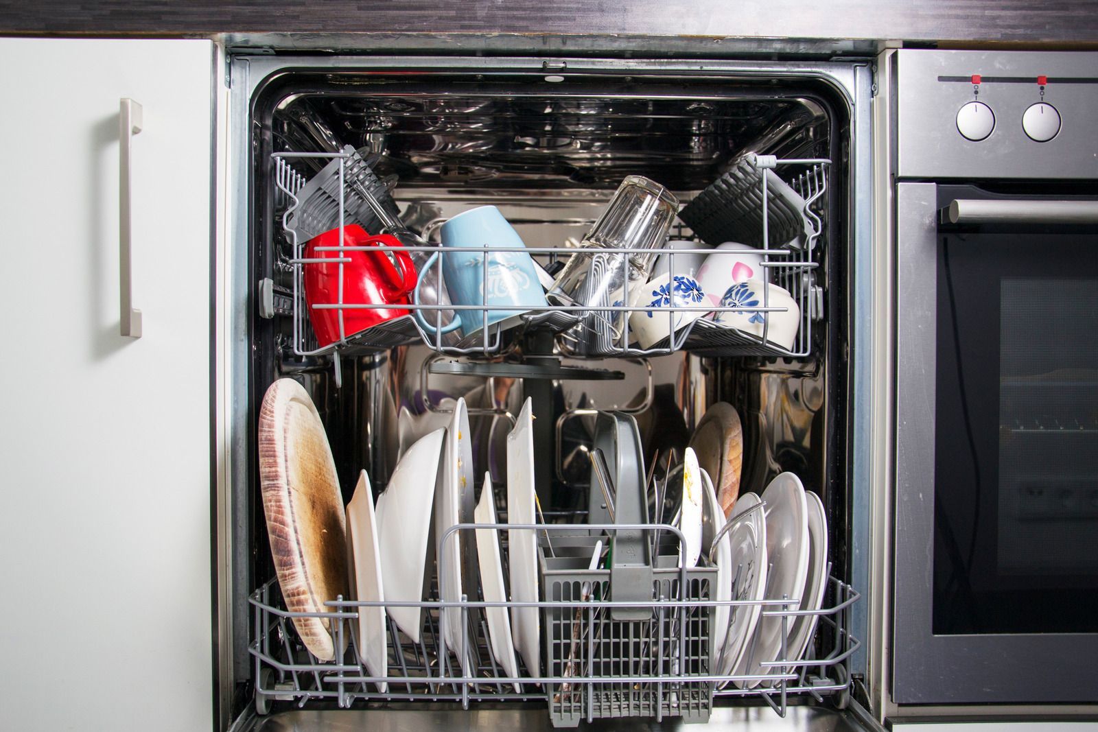 Dishwasher open, filled with dishes: plates, cups, and bowls. Stainless steel interior, kitchen setting.