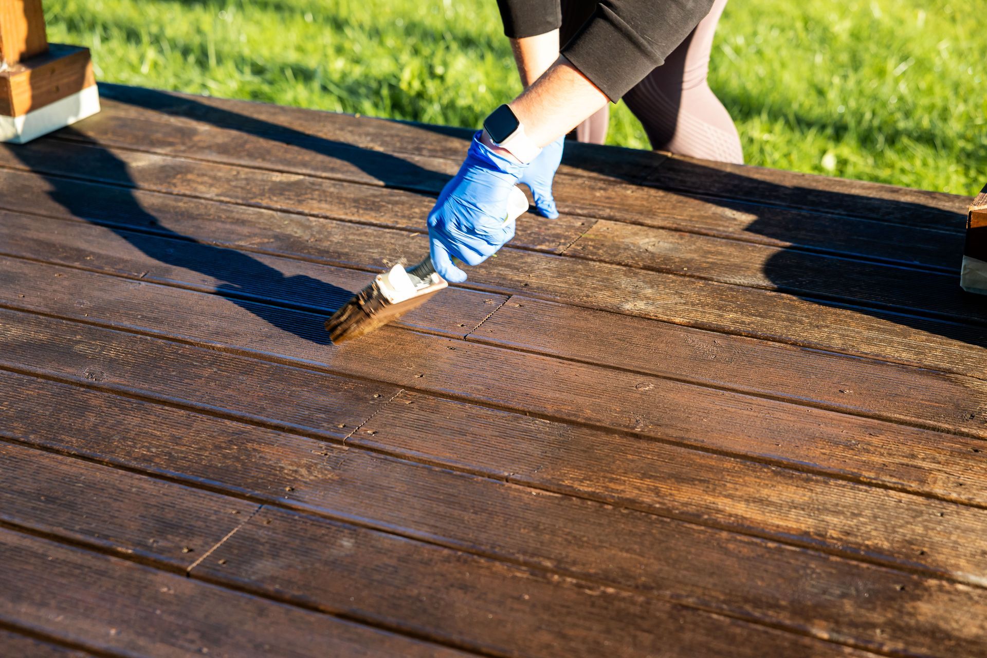 Person staining or sealing a wooden deck using a brush while wearing gloves.