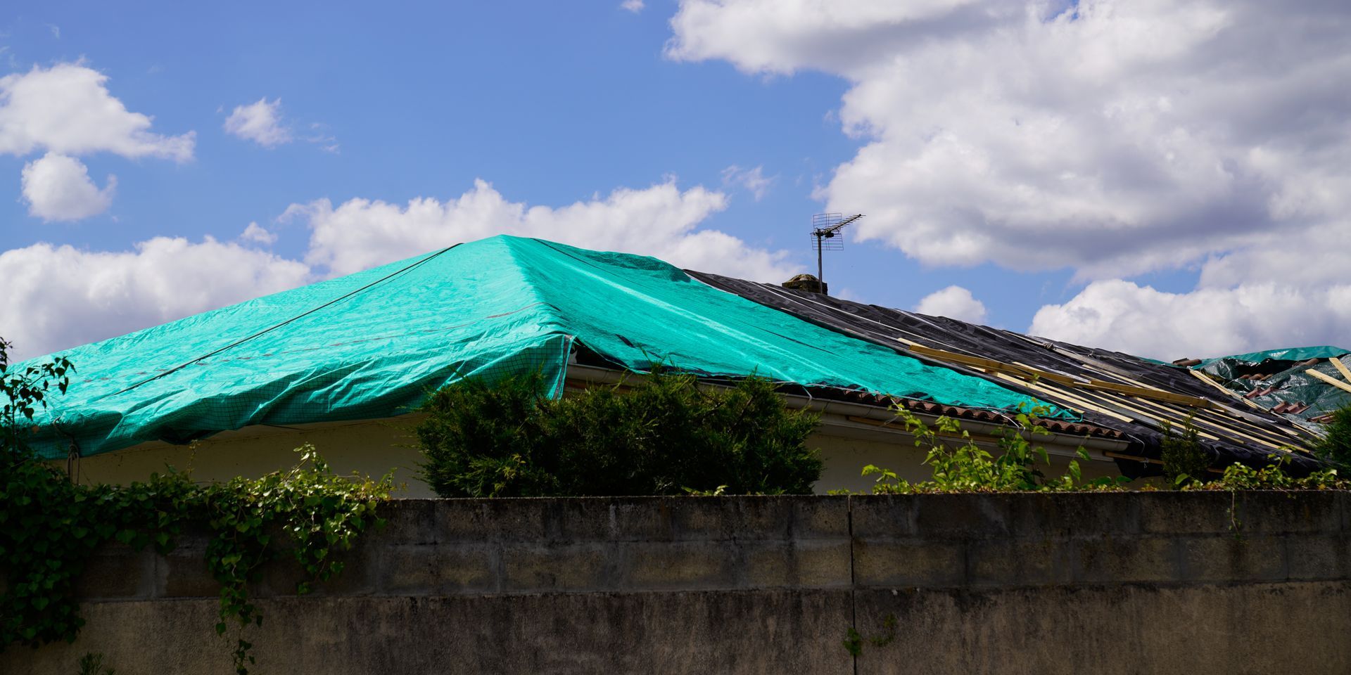 Storm-damaged house roof covered with a green plastic tarp for temporary protection.