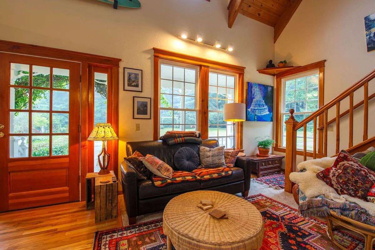 Cozy living room with dark leather sofa, colorful rugs, wooden accents, and large windows.
