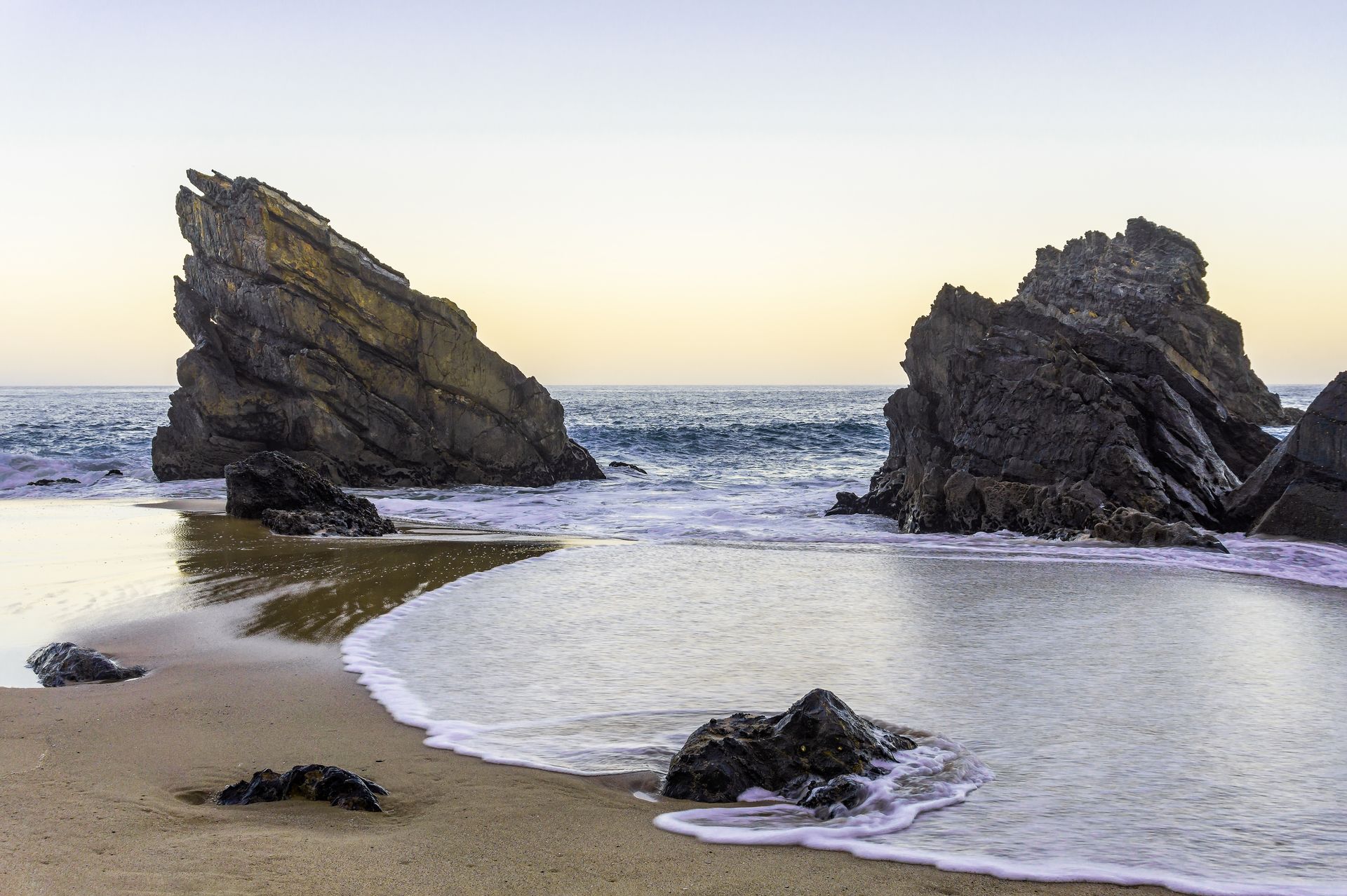 Rocky beach with waves lapping on shore, large rock formations in water, sky is light.