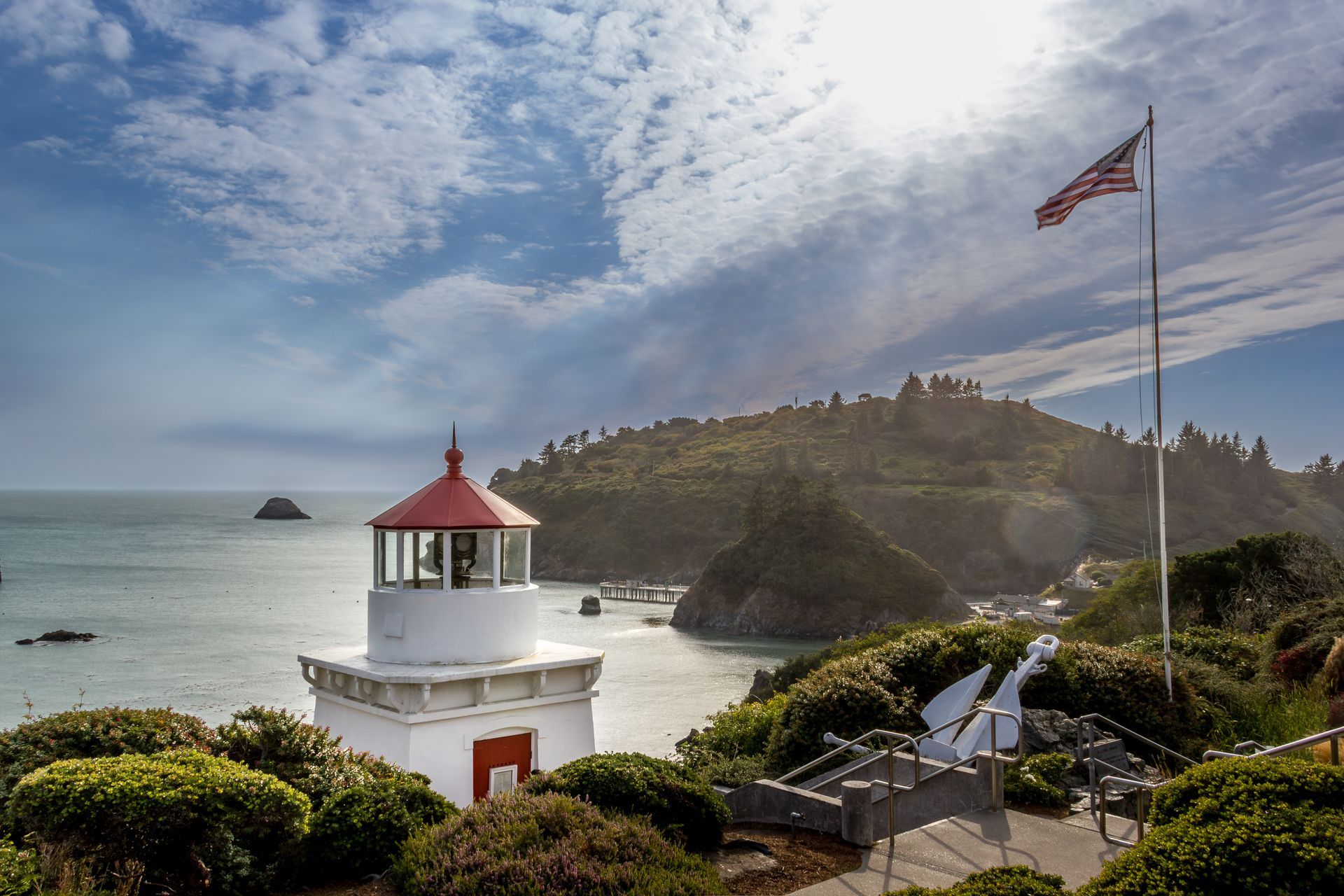 Lighthouse with red roof and American flag overlooking the ocean and coastline under a partly cloudy sky.