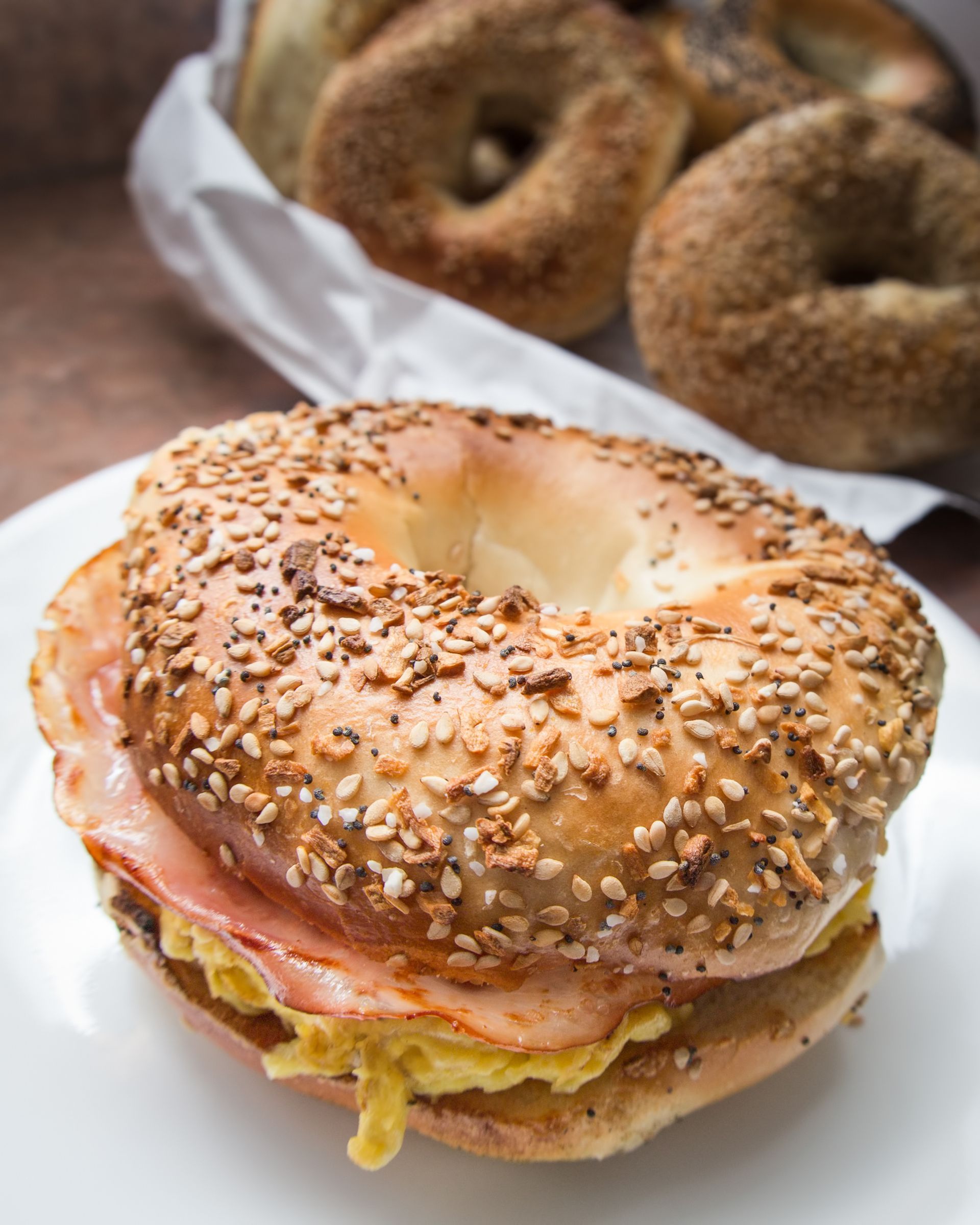 Bagel sandwich with egg and ham on a white plate; bagels in a basket in the background.