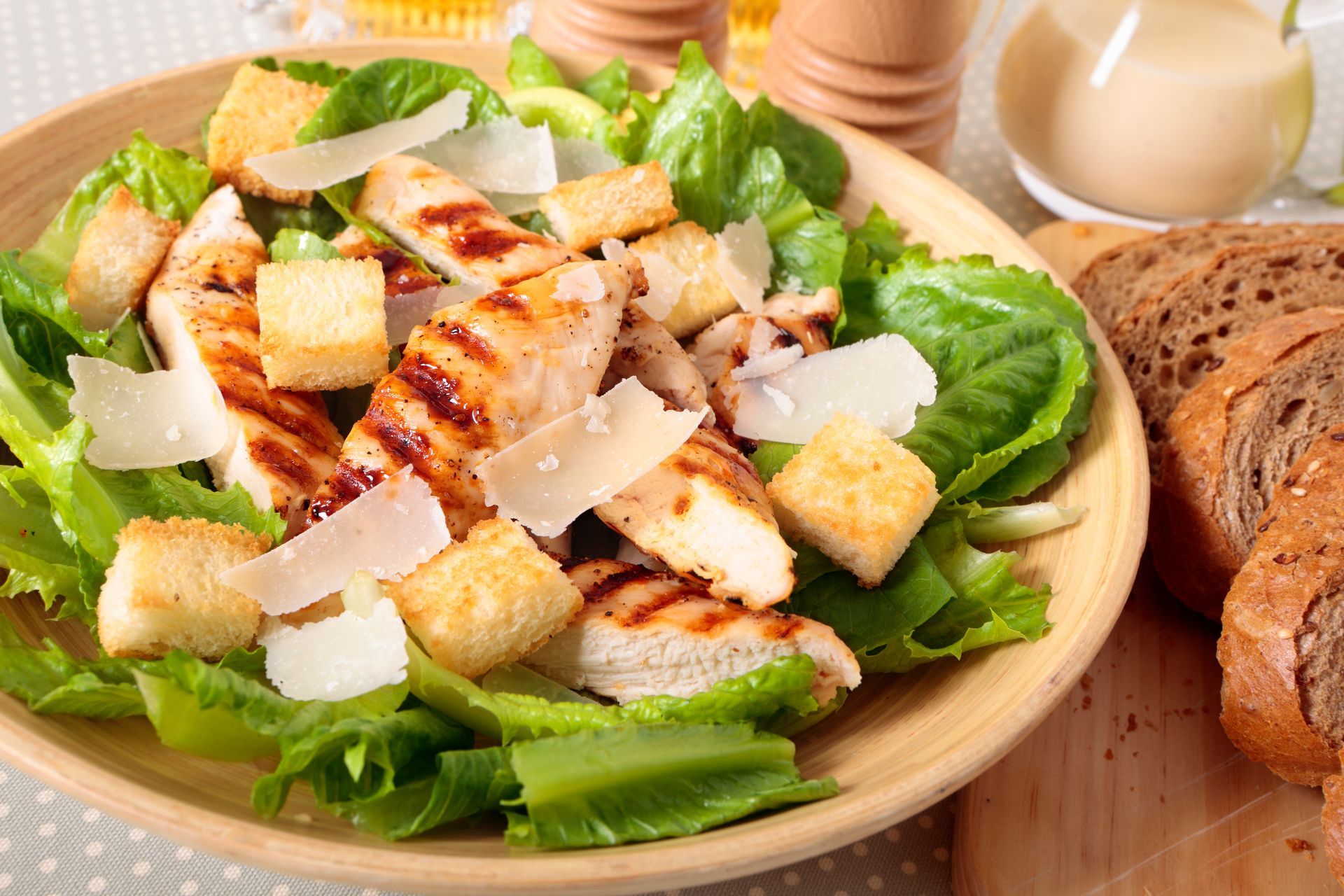 Grilled chicken Caesar salad with croutons and shaved Parmesan in a wooden bowl, bread on the side.