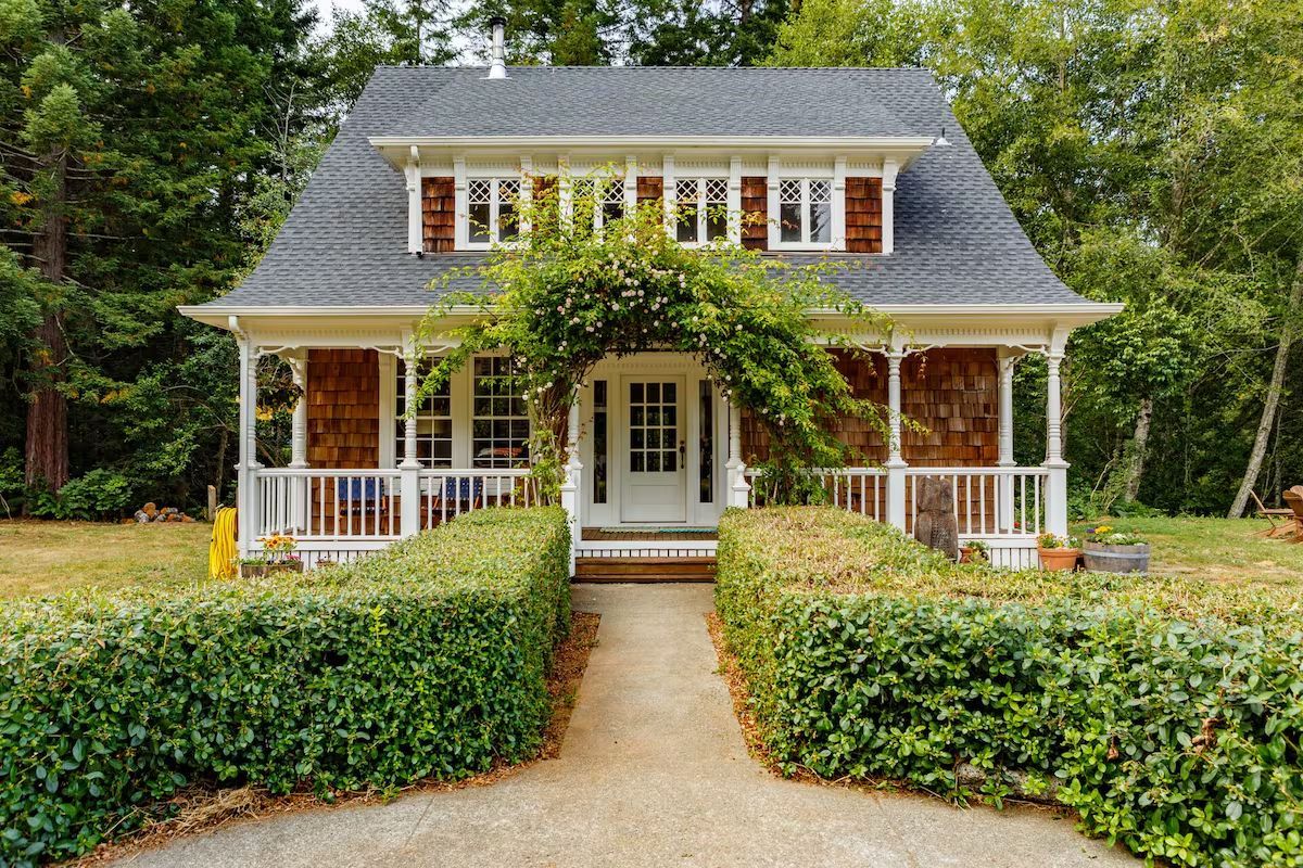 Cottage with brown shingle siding, white trim, and green hedge-lined pathway leading to the front door.