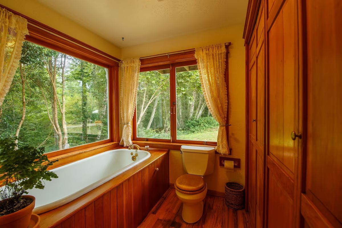 Bathroom with a large window overlooking trees, featuring a bathtub, toilet, and wooden cabinetry.