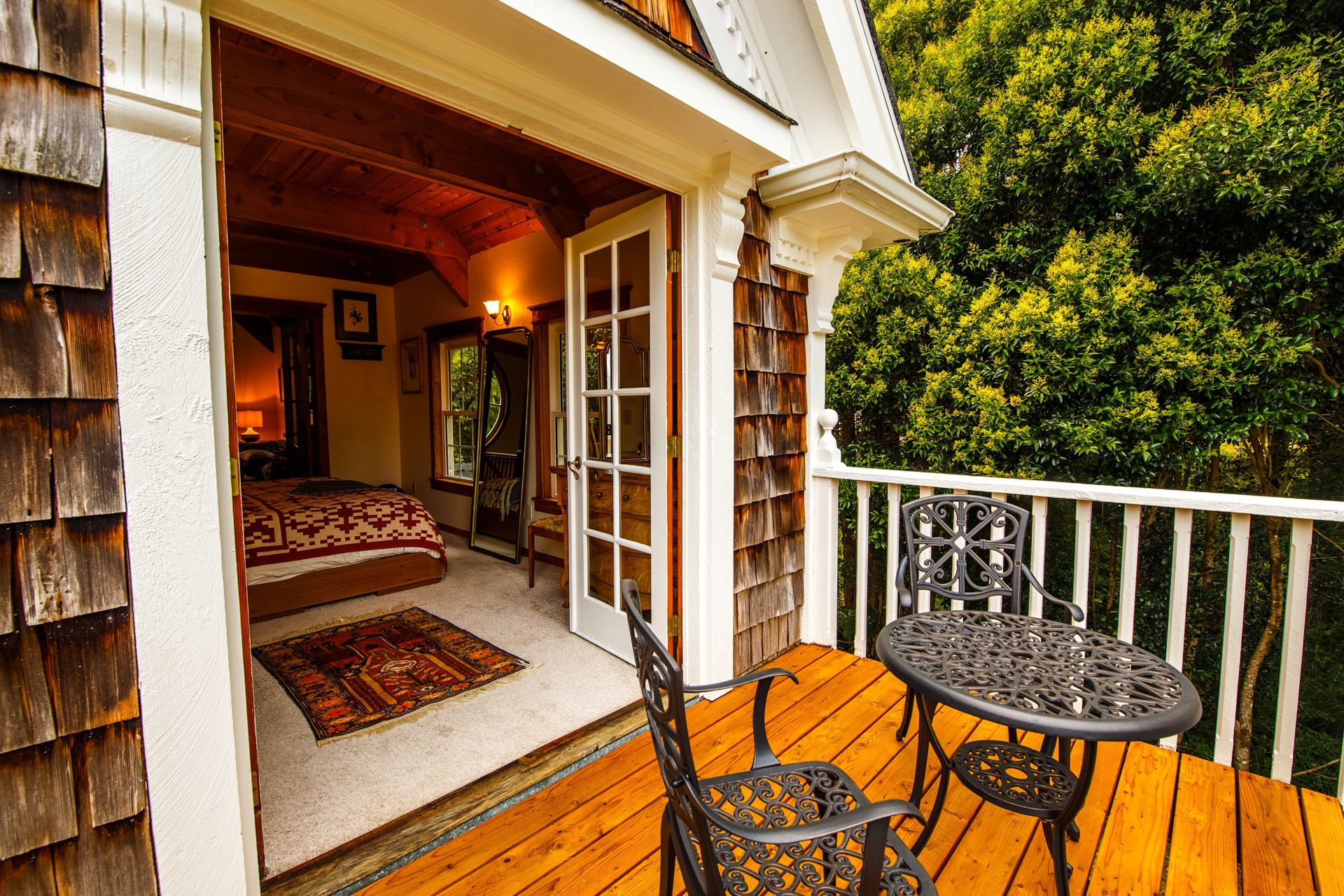 Bedroom with open doors to a small balcony with table and chairs, surrounded by trees.