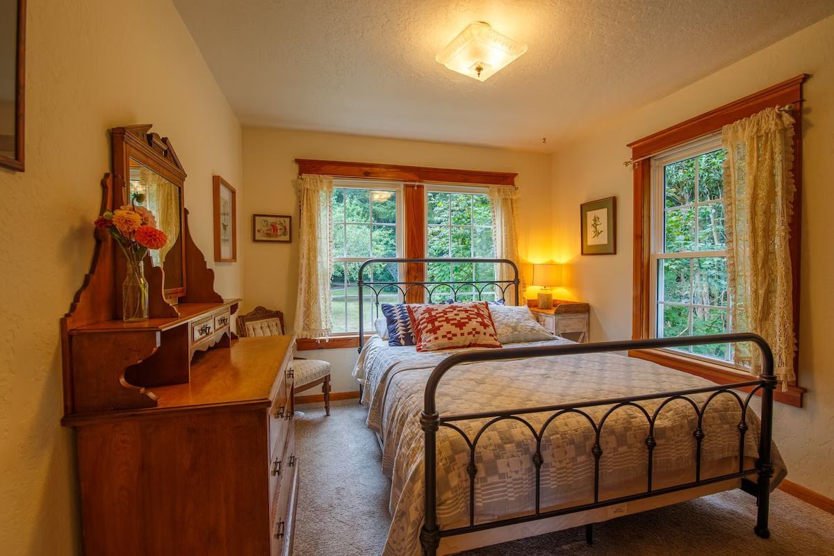 Bedroom with wood furniture, metal bed frame, windows with sheer curtains.