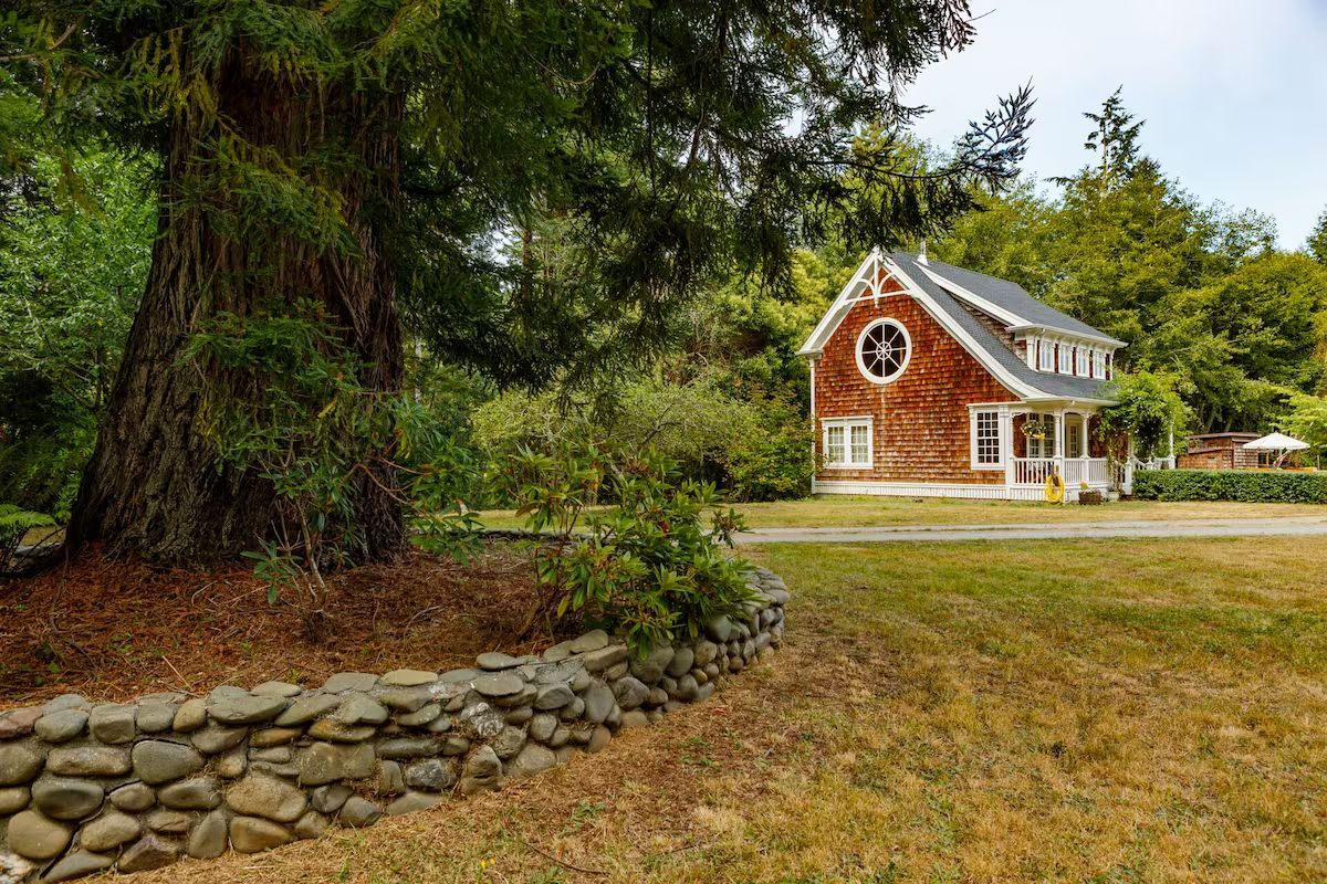 Rustic house with cedar siding, large tree, and rock wall in grassy yard.