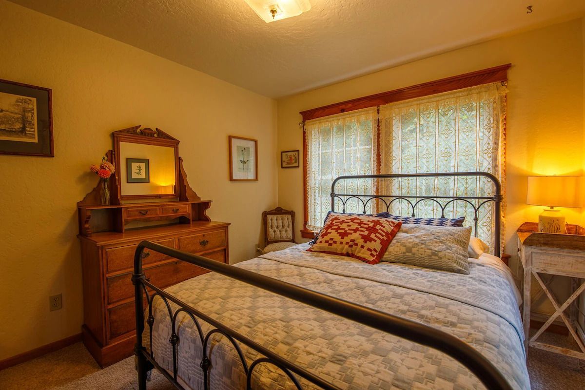 Bedroom with iron bed frame, floral bedding, antique dresser, and window with sheer curtains.