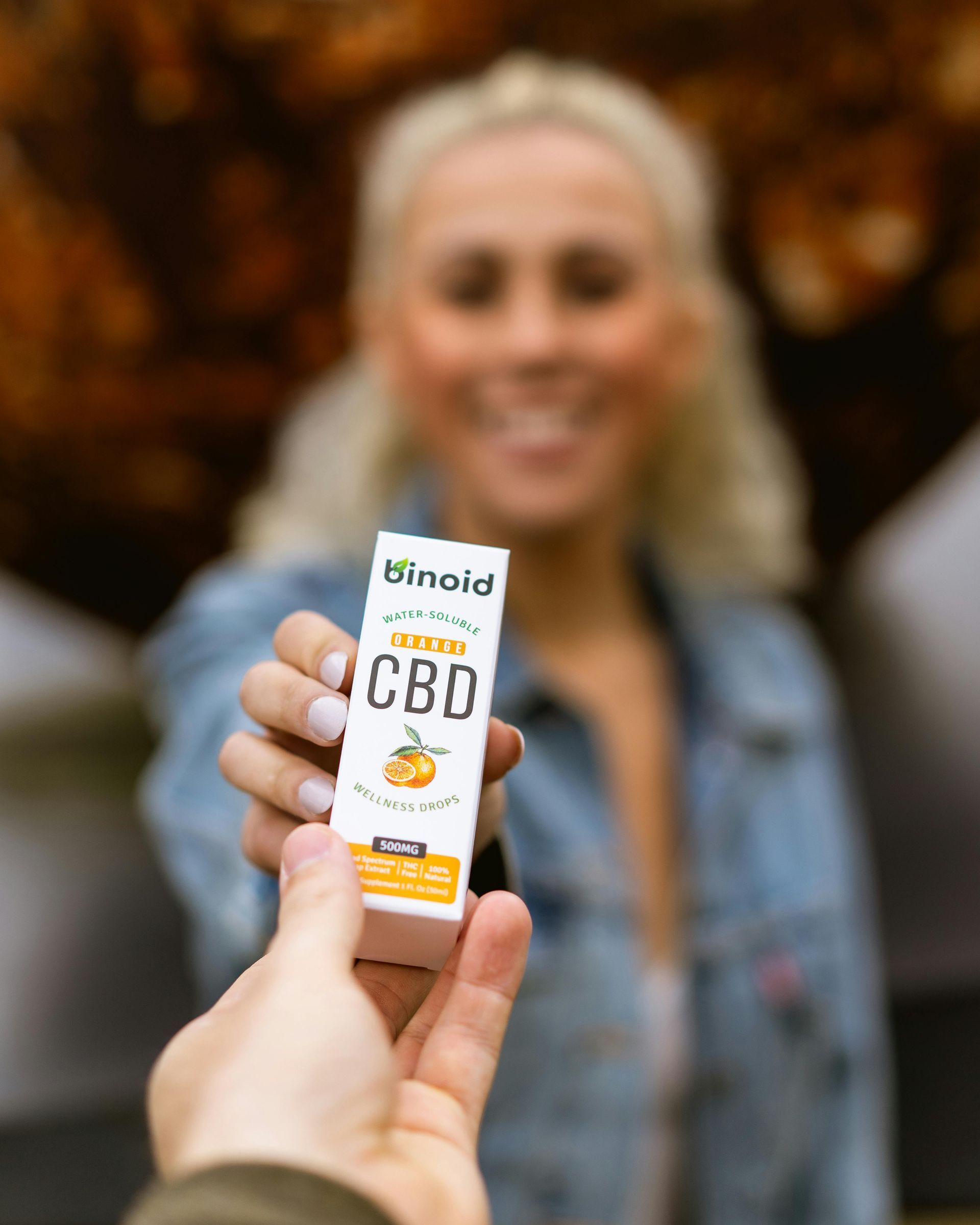 Hand holding a box of Binoid water‑soluble orange CBD wellness drops in front of a smiling customer