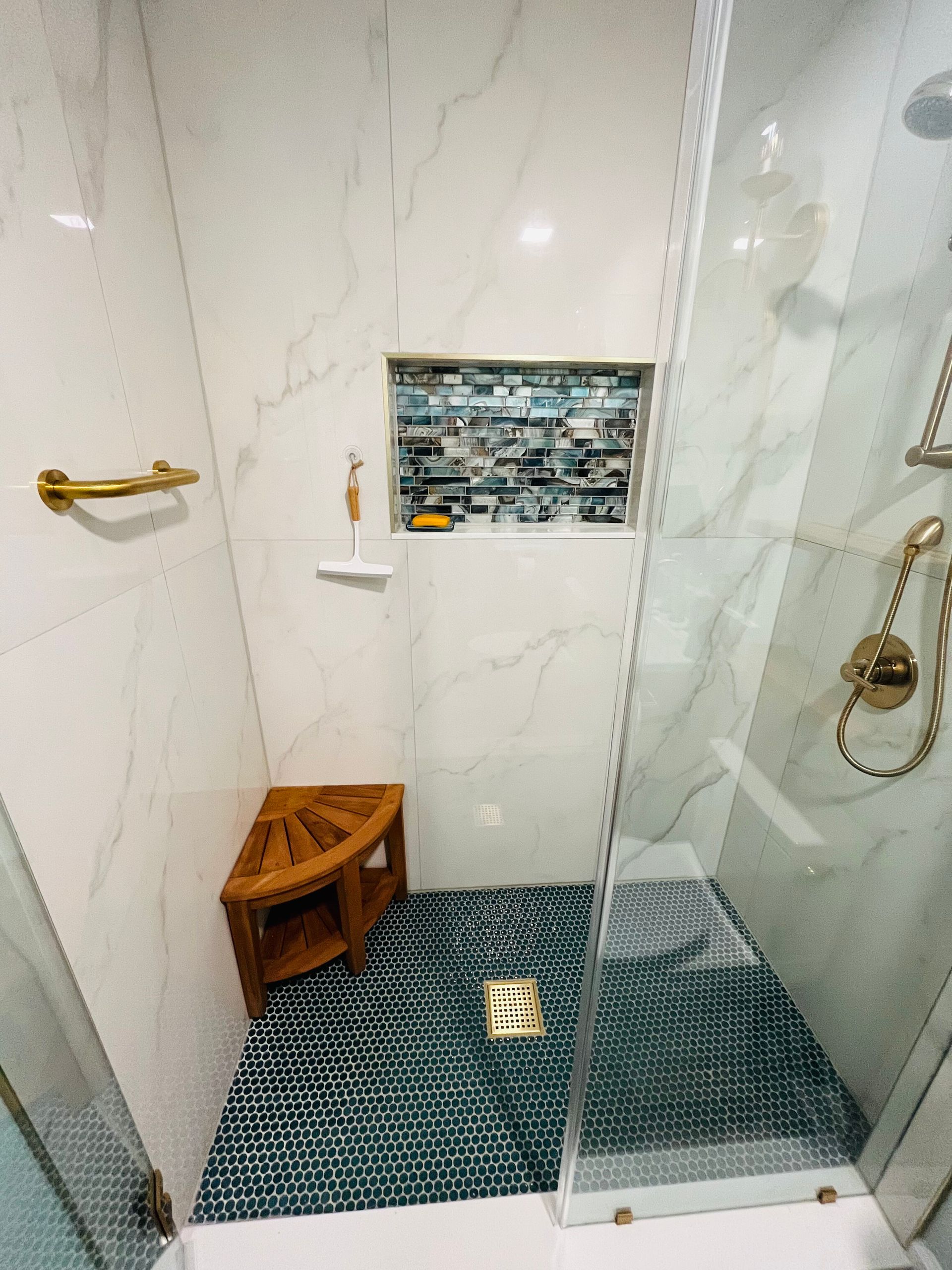 A glass-enclosed shower with white marble walls, a teal mosaic tile floor, a gold drain, a wood stool, and a recessed shelf.