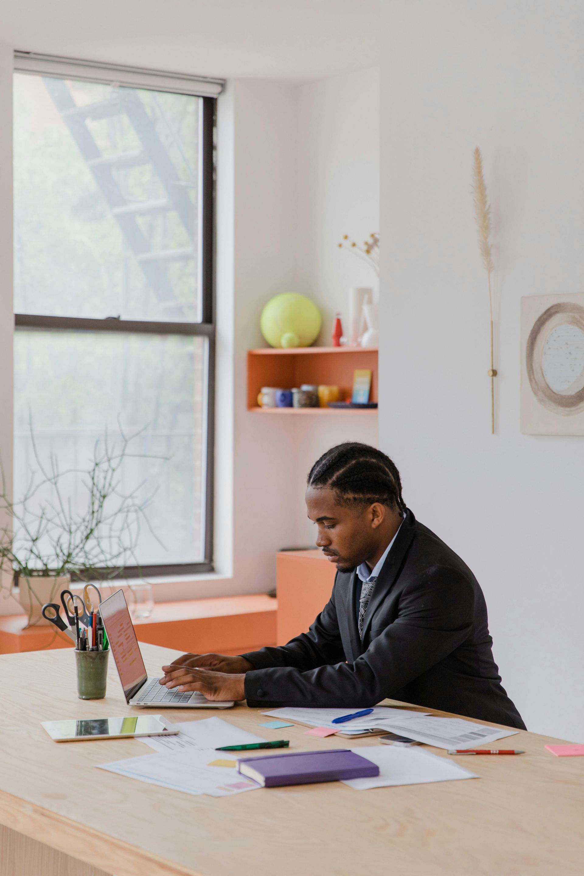 Person in a blazer typing on a laptop at a wooden desk near a window, papers and supplies are scattered about.