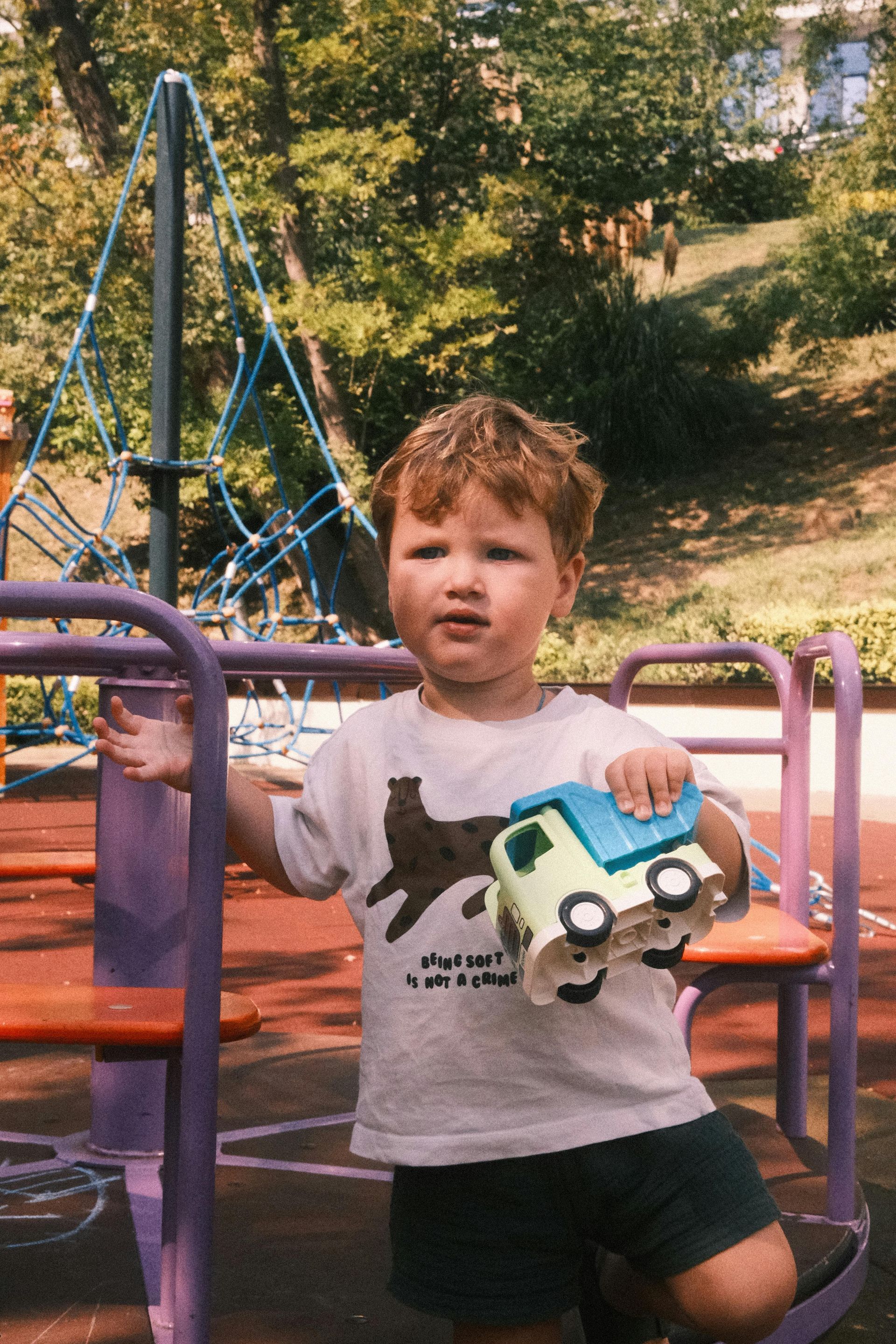 Child at playground holding toy car, standing next to purple climbing structure.