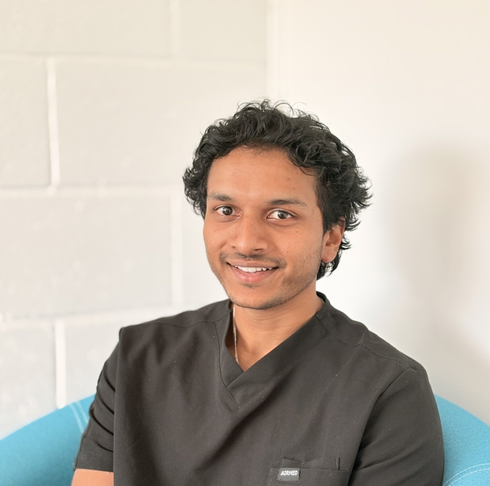 Smiling person with curly hair in dark top, seated against a white wall.