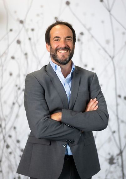 A man with a beard is wearing a suit and smiling with his arms crossed.