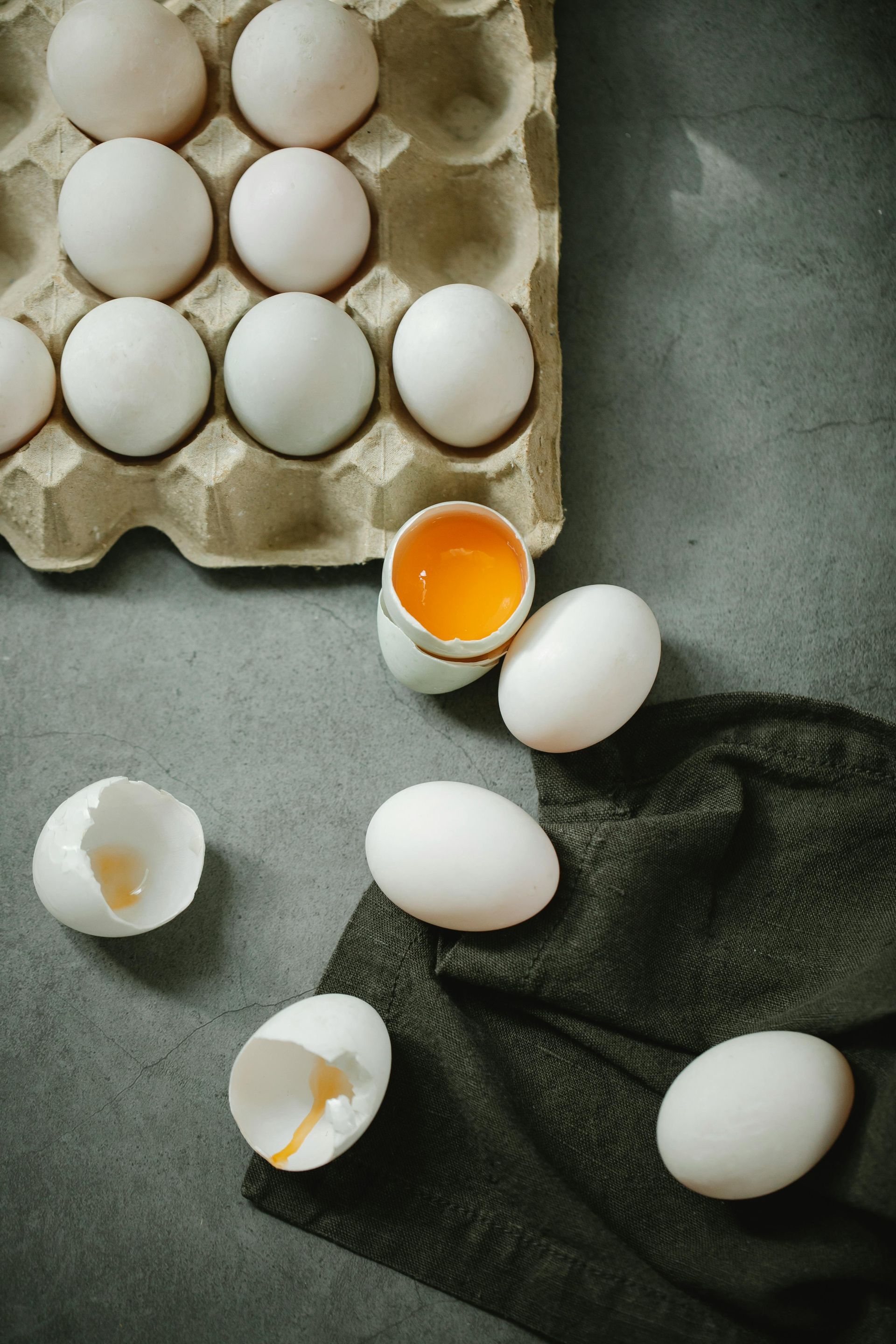 Eggs in carton, with cracked eggs and yolk spilling, on a grey surface.