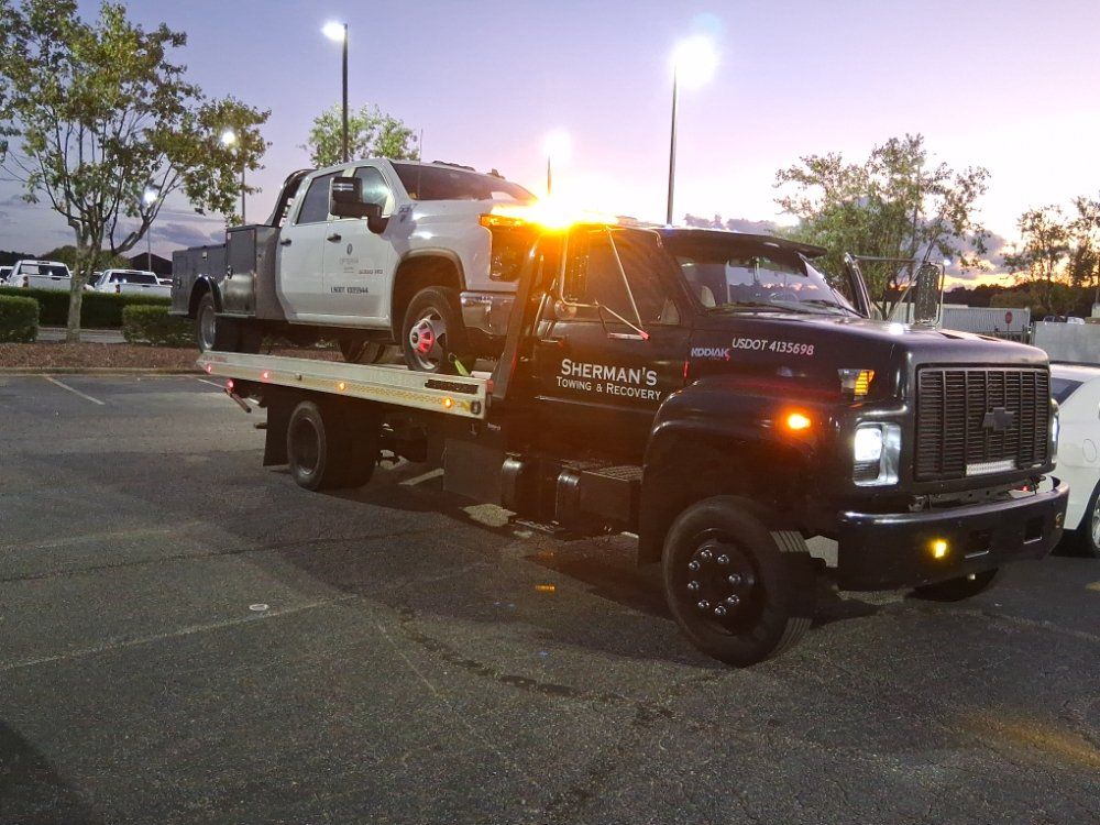 A white car being towed on a flatbed truck on a highway with a blue sky and trees in the background.