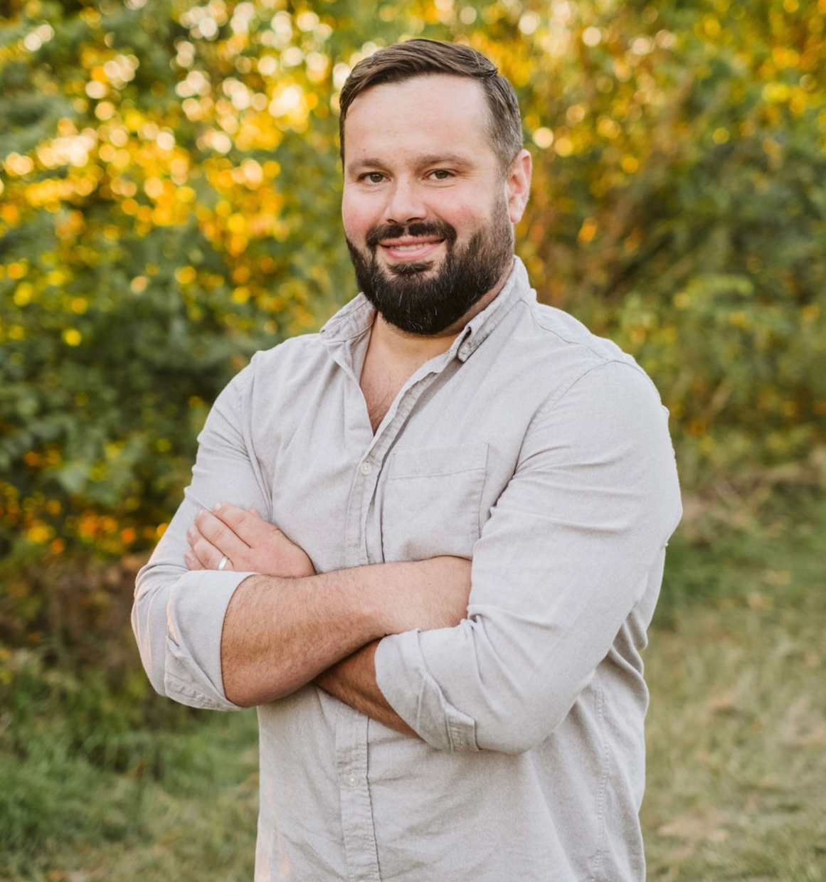 A man with a beard is standing in a field with his arms crossed.