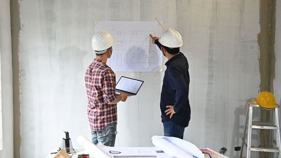 Two construction workers are looking at a blueprint on a wall.