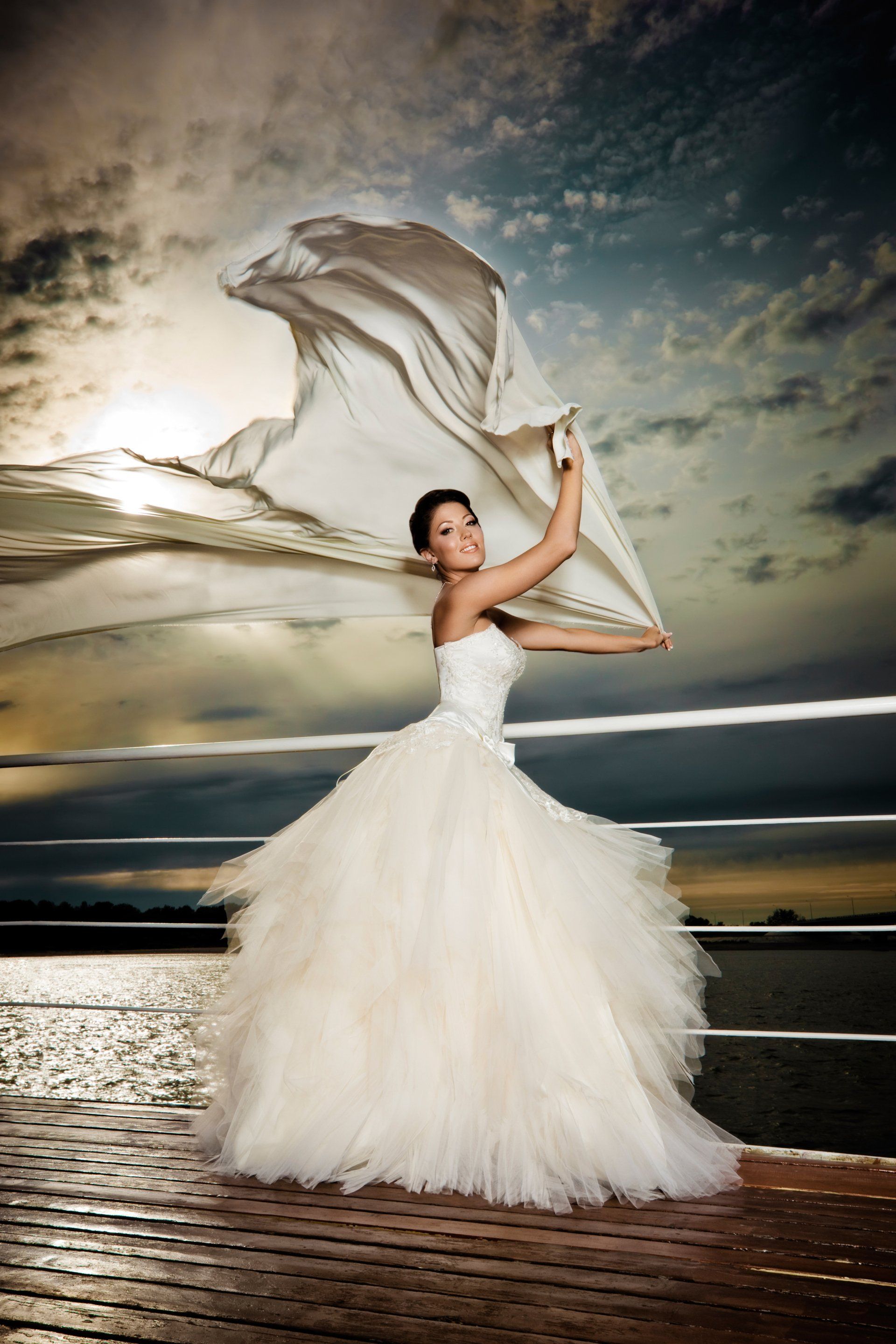 A woman in a wedding dress is standing on a pier holding a white cloth.