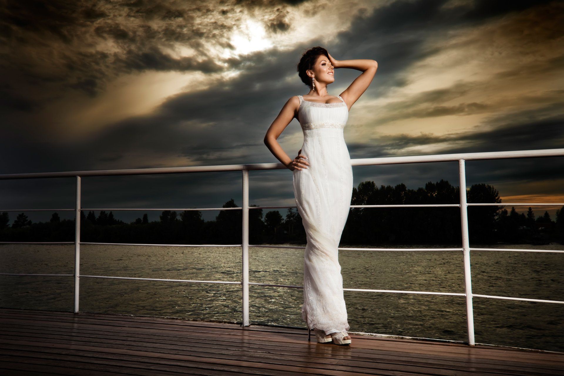 A woman in a white dress is leaning on a railing overlooking a body of water.