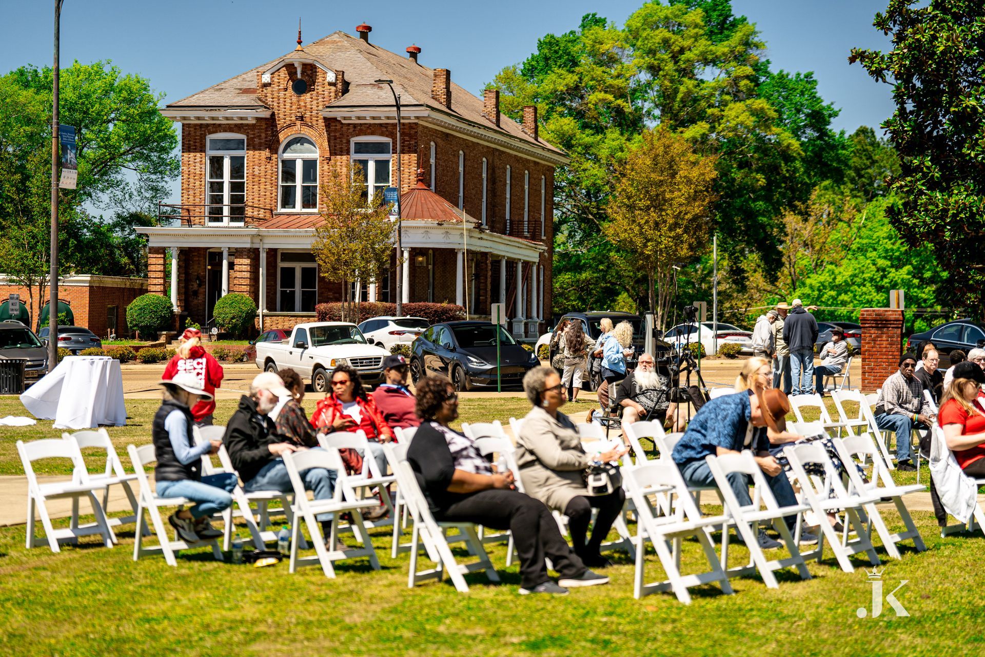 A group of people are sitting in white chairs in front of a brick building.