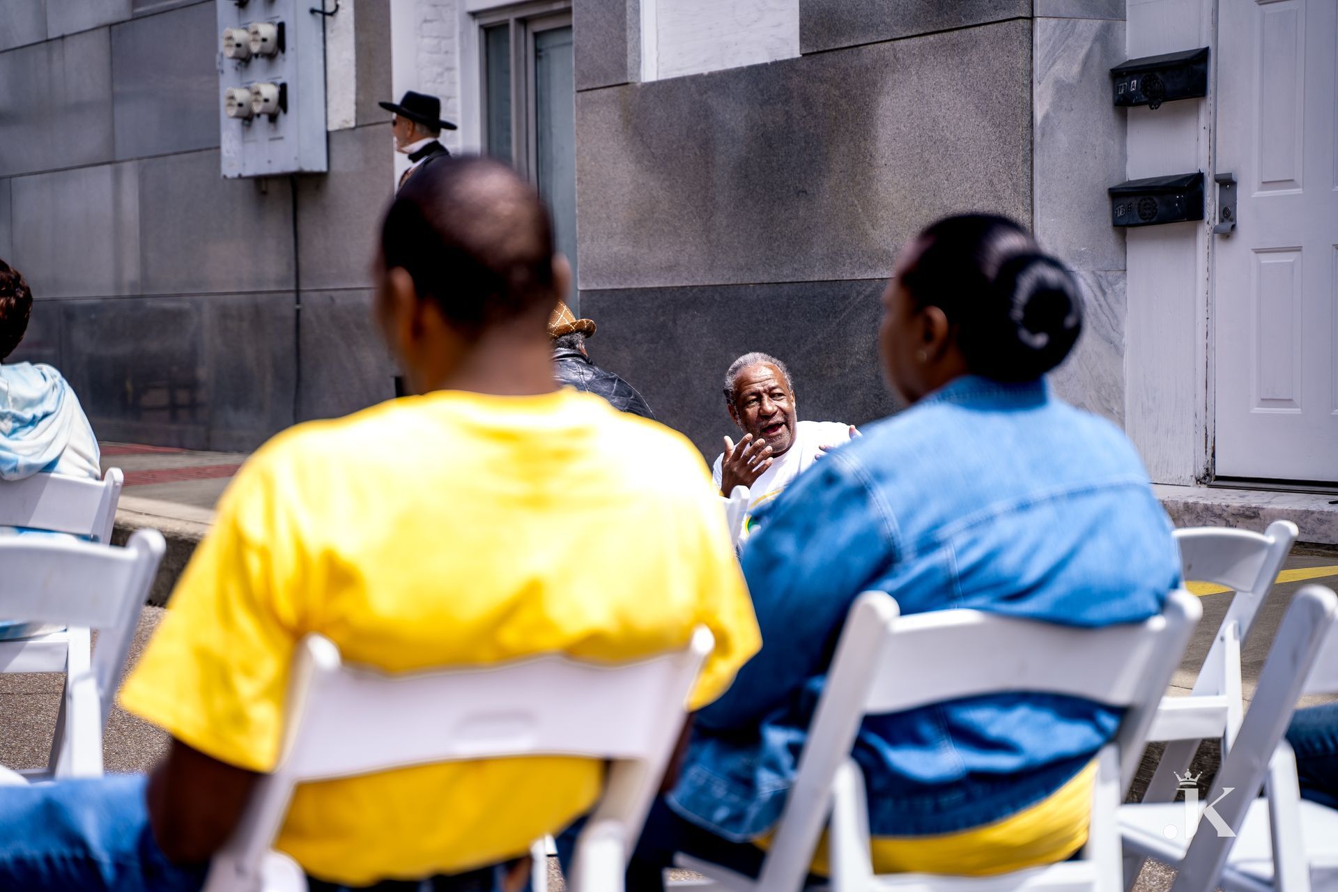 A group of people are sitting in white folding chairs outside a building.